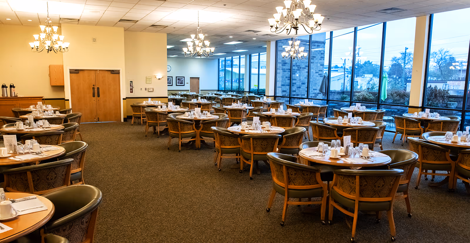 A spacious dining room with multiple round wooden tables, each surrounded by four upholstered chairs. The tables are set with white cups, glasses, and silverware. Large windows line one side of the room, allowing natural light to fill the space. Several chandeliers hang from the ceiling, providing additional lighting. The room has a carpeted floor and light-colored walls.
