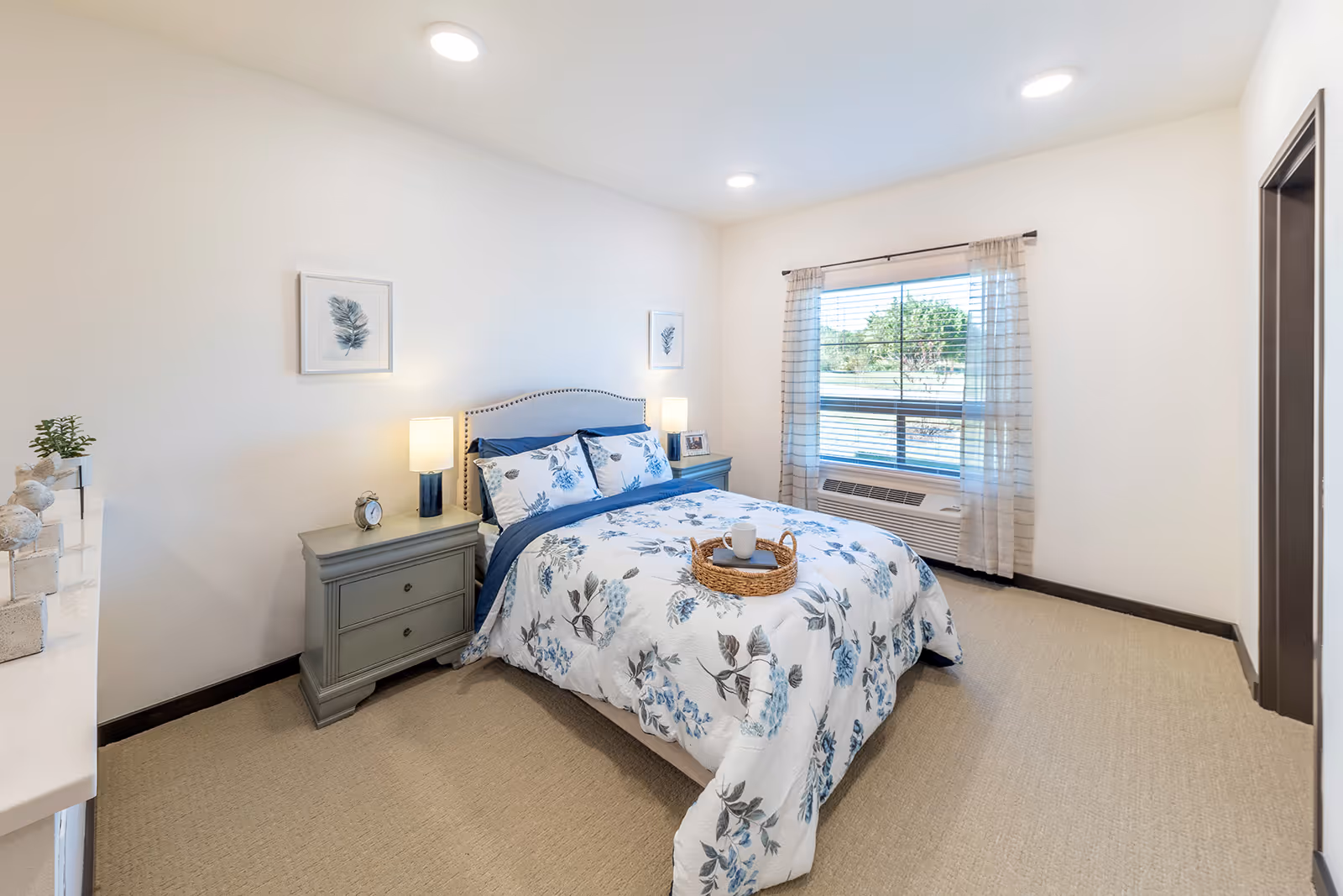 A bright and cozy bedroom with a bed dressed in white and blue floral bedding. The bed has a beige upholstered headboard and is flanked by two gray nightstands, each with a lamp. There are two framed botanical prints on the wall above the nightstands. A window with sheer curtains lets in natural light, and a beige carpet covers the floor.