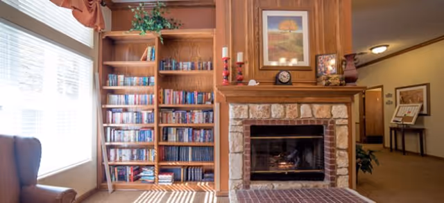 A cozy living room area featuring a stone fireplace with a wooden mantel decorated with candles, a clock, and framed pictures. To the left of the fireplace is a tall wooden bookshelf filled with books and a small plant on top. A large window with blinds allows natural light to fill the room, and a comfortable armchair is partially visible on the left side.
