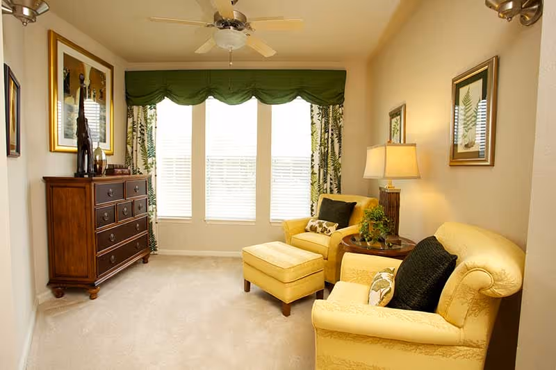 A cozy living room with two yellow upholstered armchairs, one with an ottoman, a wooden side table with a lamp and plant, a wooden dresser with decorative items, three large windows with green valance and patterned curtains, and framed artwork on the walls.