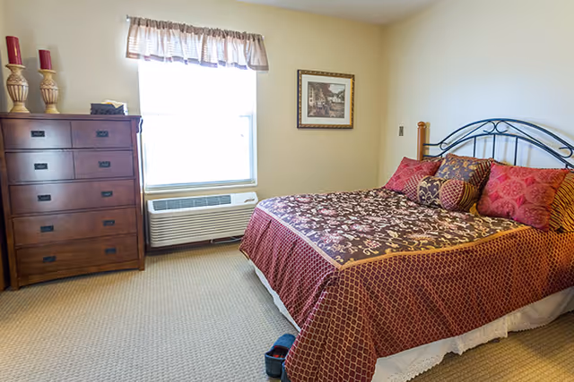 A bedroom with a bed covered in a patterned red and brown bedspread with multiple pillows. There is a wooden dresser with three red candles on top to the left of a window with a valance. Below the window is an air conditioning unit. A framed picture hangs on the wall above the bed.