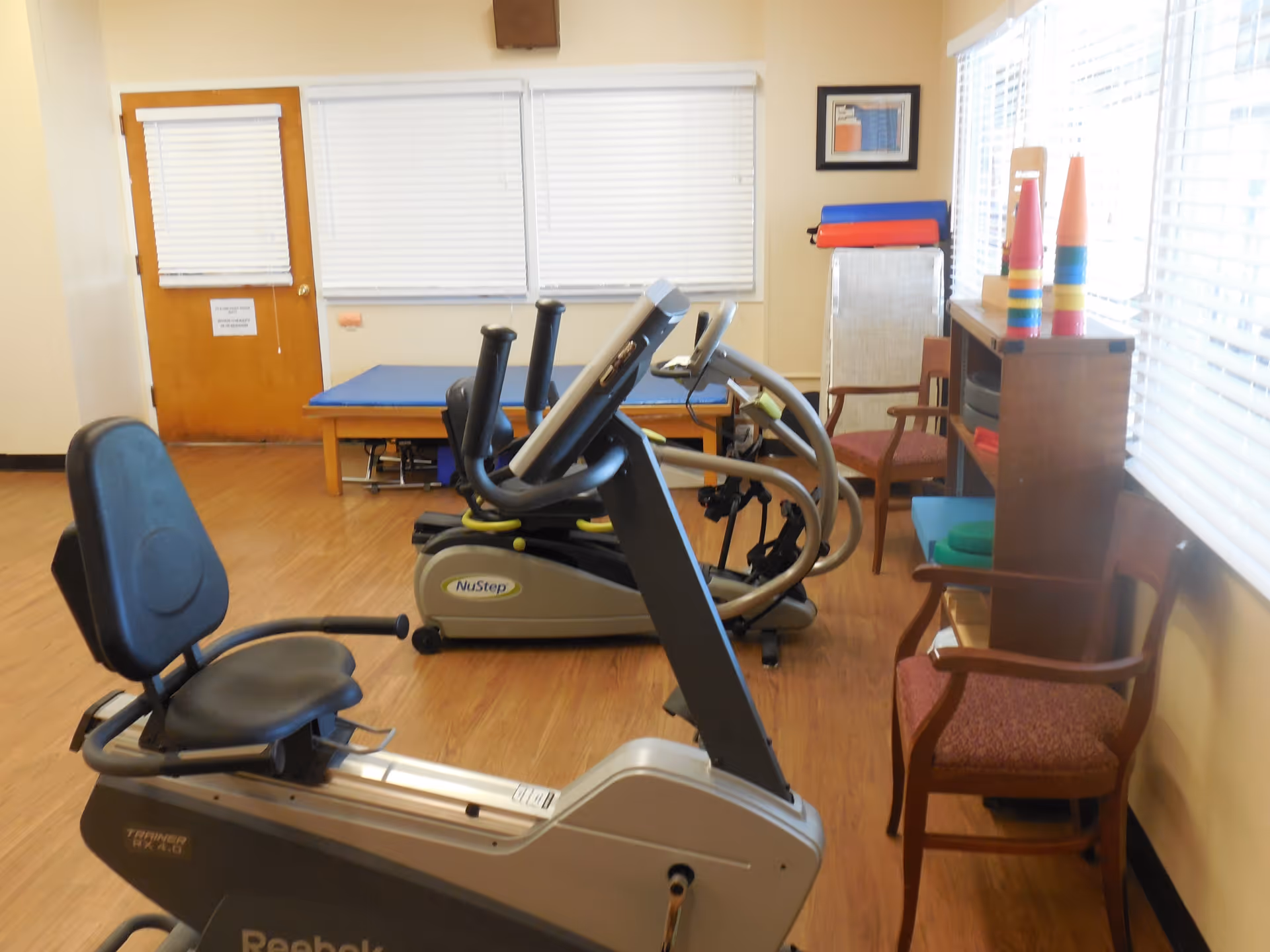 A small exercise room with wooden flooring containing a recumbent exercise bike and two NuStep machines. There are two wooden chairs with red cushions, a wooden shelf with colorful cones and exercise mats, and a padded table against the wall. The room has windows with white blinds and a wooden door.