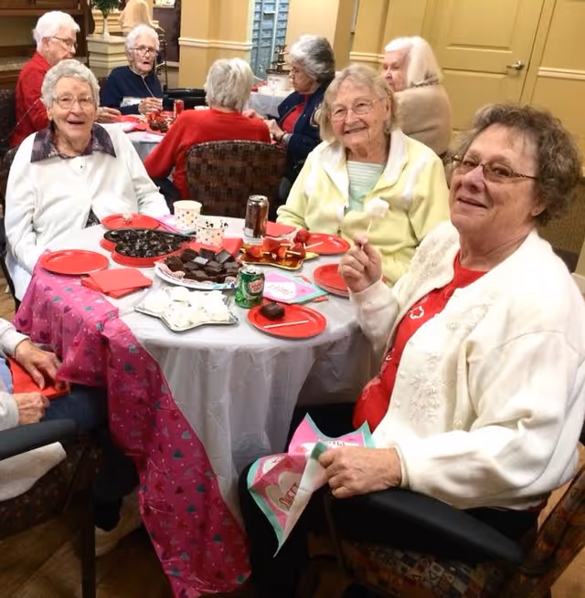 Several elderly women smiling and seated around a table with plates of desserts and drinks in a communal dining room.