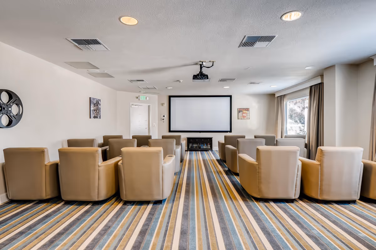 A small theater room with two rows of beige armchairs facing a large blank projector screen mounted on the wall. The room has a striped carpet in shades of blue, gray, and beige, and a window with curtains on the right side letting in natural light. There are movie-themed decorations on the walls and ceiling lights.