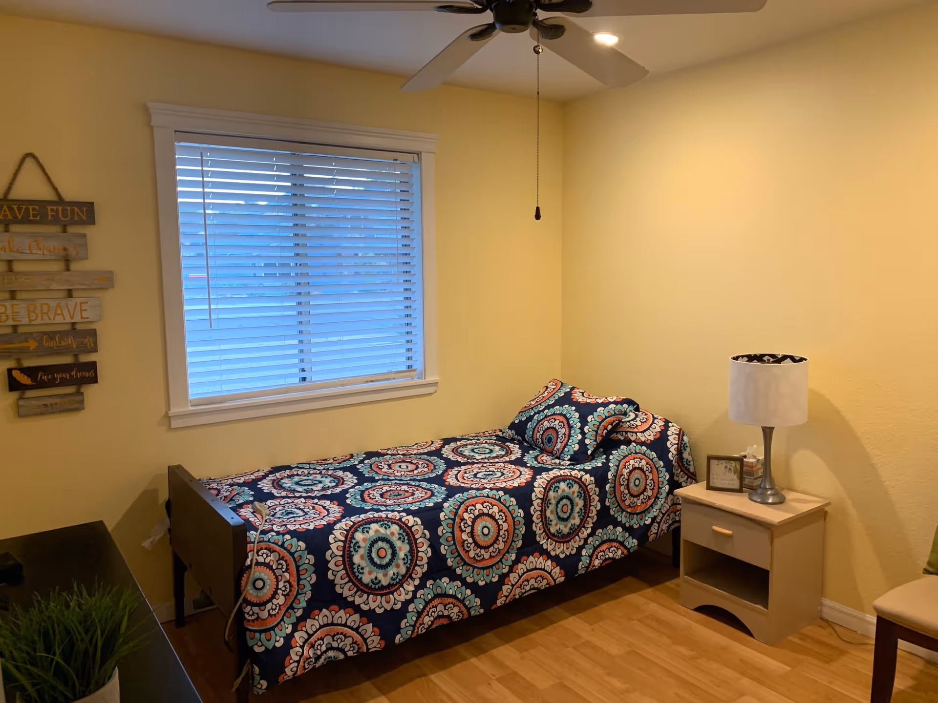 A small bedroom with a single bed covered in a colorful patterned bedspread and matching pillow. There is a window with white blinds above the bed, a ceiling fan with a light, a nightstand with a lamp and a small framed picture, and a decorative wall hanging with inspirational words on the left wall.