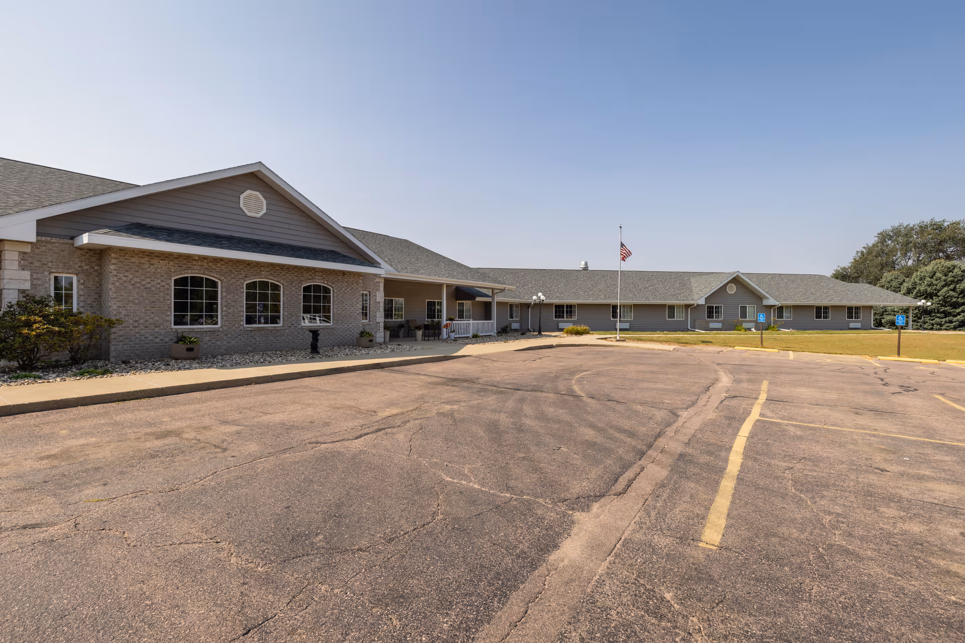 Single-story brick and siding senior living building with a covered front entrance, flagpole, and empty parking lot under a clear sky.
