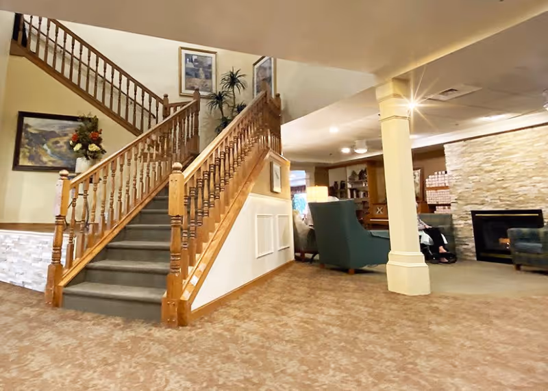 Interior view of a senior living facility showing a carpeted floor, a wooden staircase with railings, a stone fireplace, and comfortable seating including armchairs. The walls are decorated with framed artwork and there is a potted plant near the staircase.