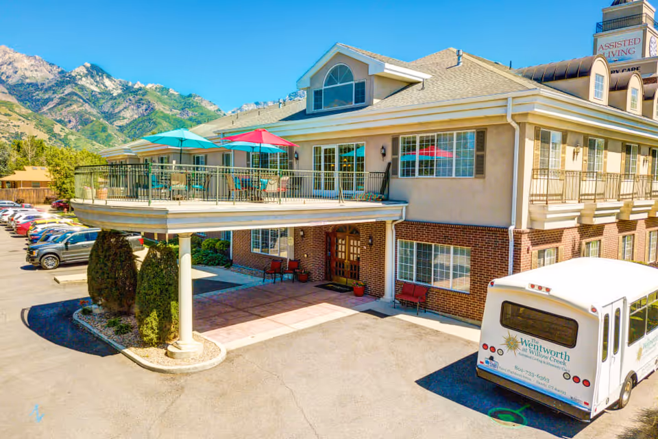 Front exterior of a two-story assisted living building with a covered entrance, second-floor balcony with colorful umbrellas, a parked shuttle bus, and mountains in the background.