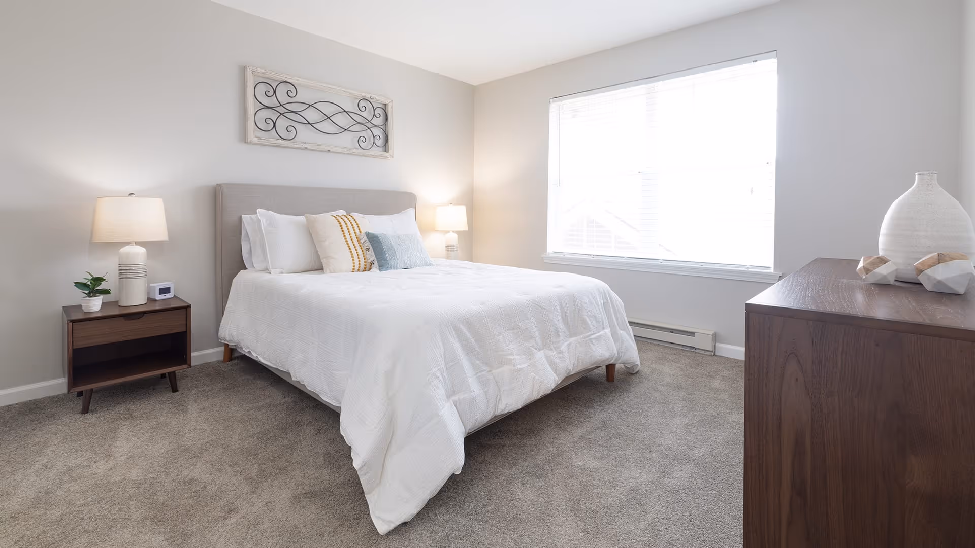 A bright and clean bedroom with a large bed covered in white bedding and multiple pillows. There are two bedside tables with lamps on each side of the bed. A decorative wall piece hangs above the bed. A large window with blinds lets in natural light, and a wooden dresser with decorative items is positioned on the right side of the room. The floor is carpeted in a neutral tone.