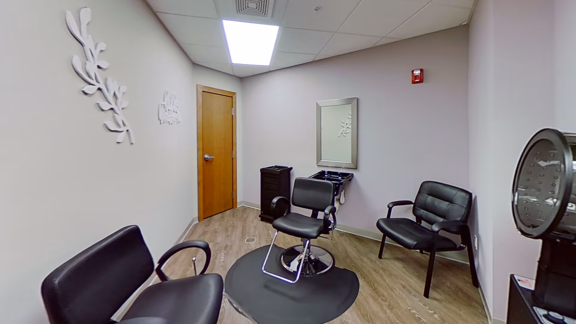 A small salon room with a black salon chair on a circular mat in the center, two black waiting chairs, a wall-mounted mirror above a small black sink, a wooden door, and light-colored walls with decorative wall art.
