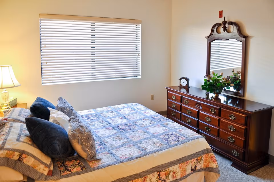 A bedroom with a bed covered in a patchwork quilt and several pillows. Next to the bed is a nightstand with a lamp. Across from the bed is a wooden dresser with multiple drawers, a decorative clock, a vase with flowers, and a large mirror mounted on the wall above it. A window with closed blinds is on the wall behind the bed.