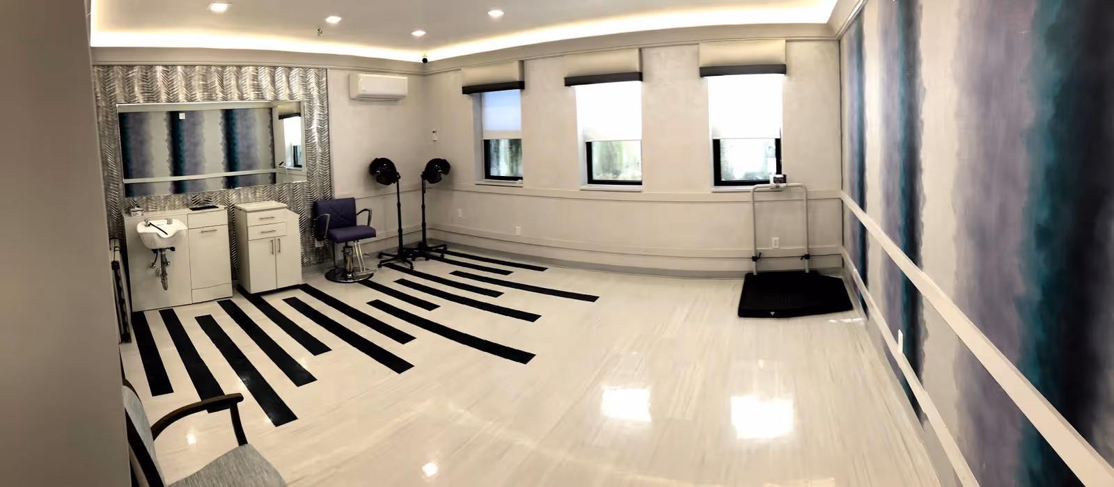 Bright rehabilitation salon with hair-styling stations, chairs and hooded dryers, a mirrored sink area, windows and handrails along a patterned wall.