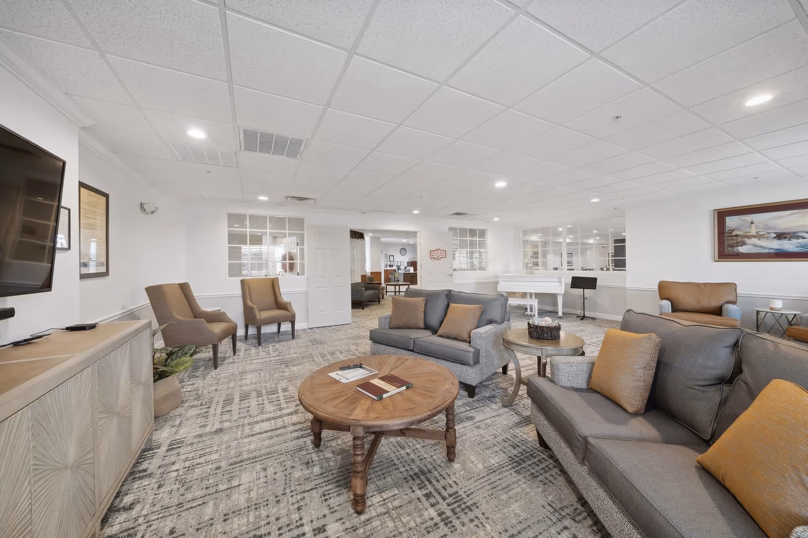 A spacious and well-lit living room area with gray sofas and orange cushions, a round wooden coffee table with books on it, two brown armchairs, a white piano in the background, a flat-screen TV mounted on the wall, and a framed painting of a lighthouse on the wall.