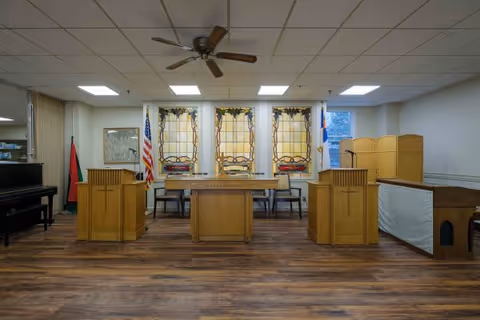 Interior view of a chapel or worship room with wooden podiums and a central altar table. Behind the altar are three stained glass windows with religious designs. There are American and Christian flags on the left side, a piano on the far left, and a wooden folding screen on the right. The room has a ceiling fan and recessed lighting.