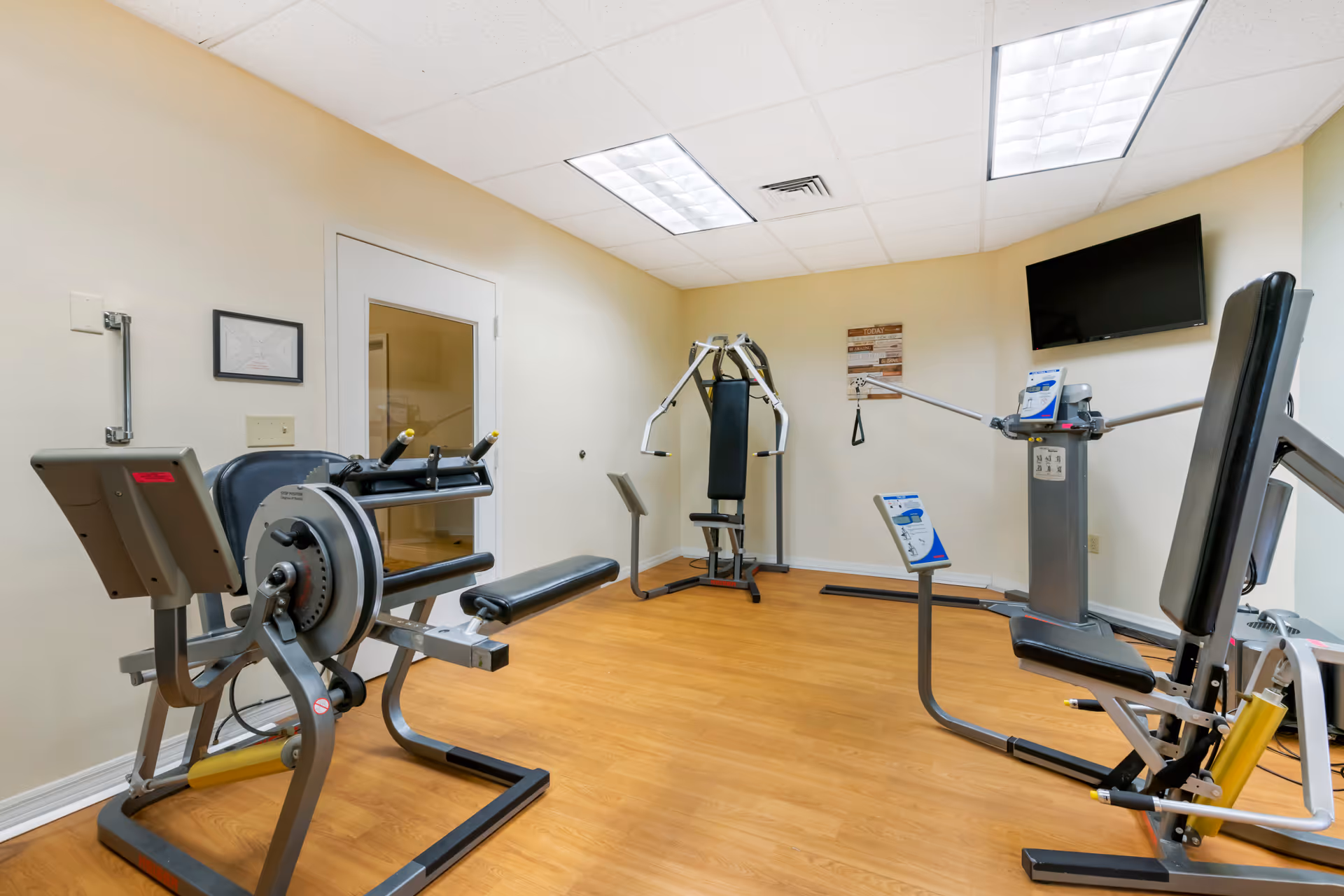 A small fitness room with exercise equipment including a rowing machine, a multi-function weight machine, and a seated chest press. The room has light-colored walls, a wooden floor, a white door with a window, and a wall-mounted flat screen TV.