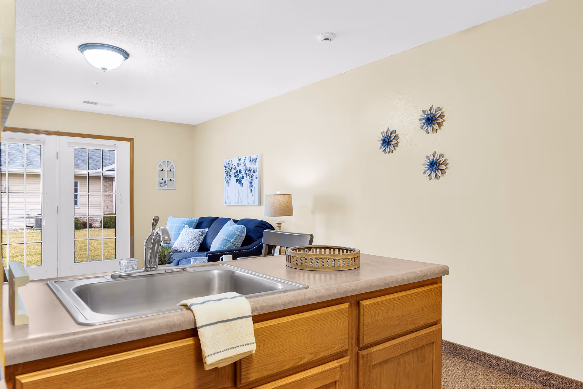View of a kitchen sink with a beige towel hanging over the edge of the counter. In the background, there is a living room area with a blue couch, decorative pillows, a lamp, and wall art featuring blue flowers. French doors lead to an outdoor area with grass and neighboring buildings.