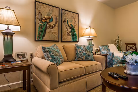 Cozy living room with a beige loveseat, teal patterned pillows and chair, peacock artwork, lamps, and a coffee table.