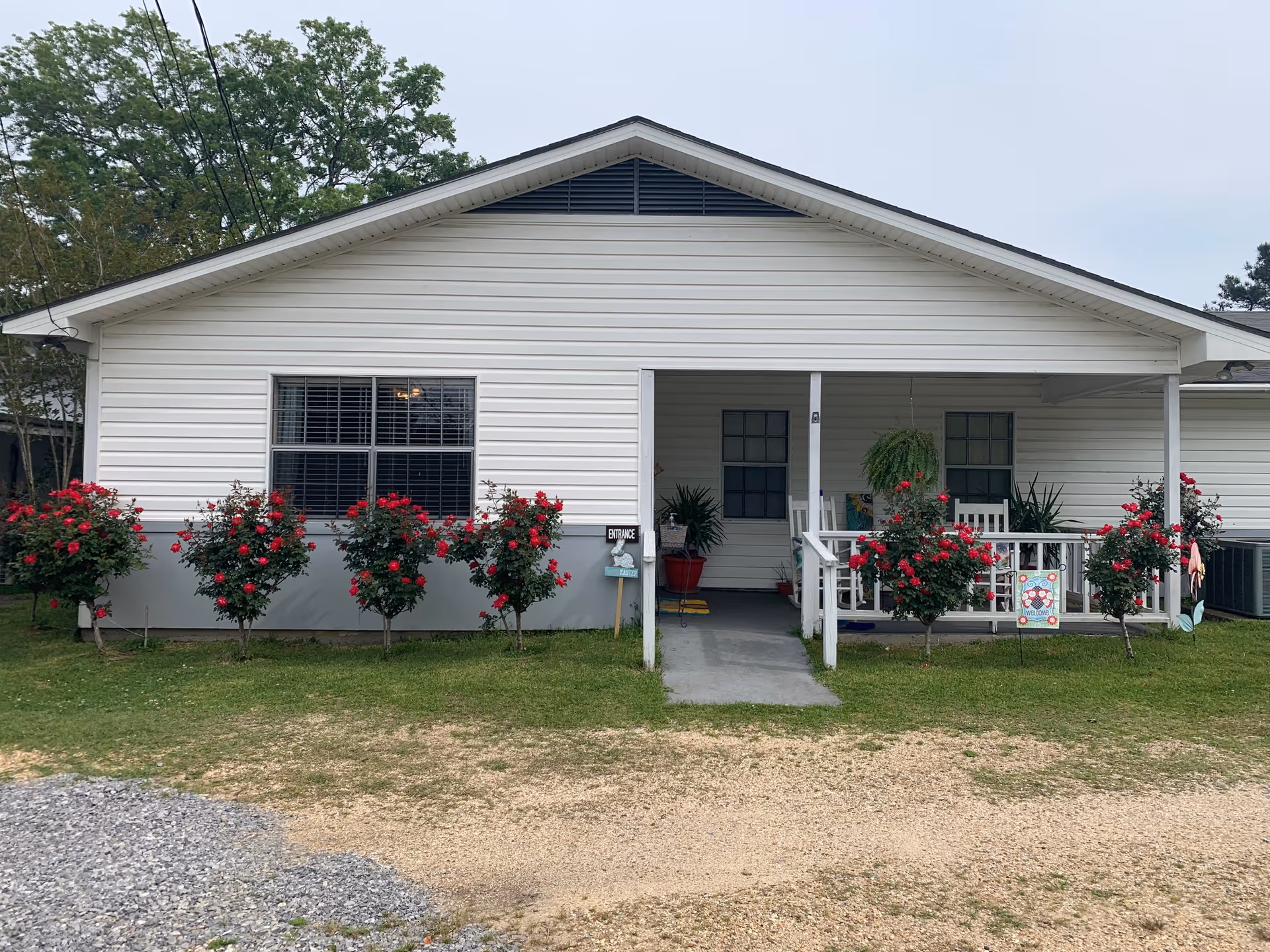 Front exterior view of a single-story white building with a small porch. There are six rose bushes with red flowers planted in front of the building. The porch has white railings, a hanging plant, and some potted plants. A sign near the entrance reads 'ENTRANCE'.