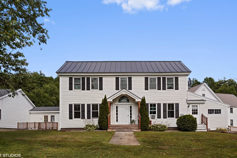 Two-story white building with black shutters and a central front entrance on a grassy lawn under a blue sky.