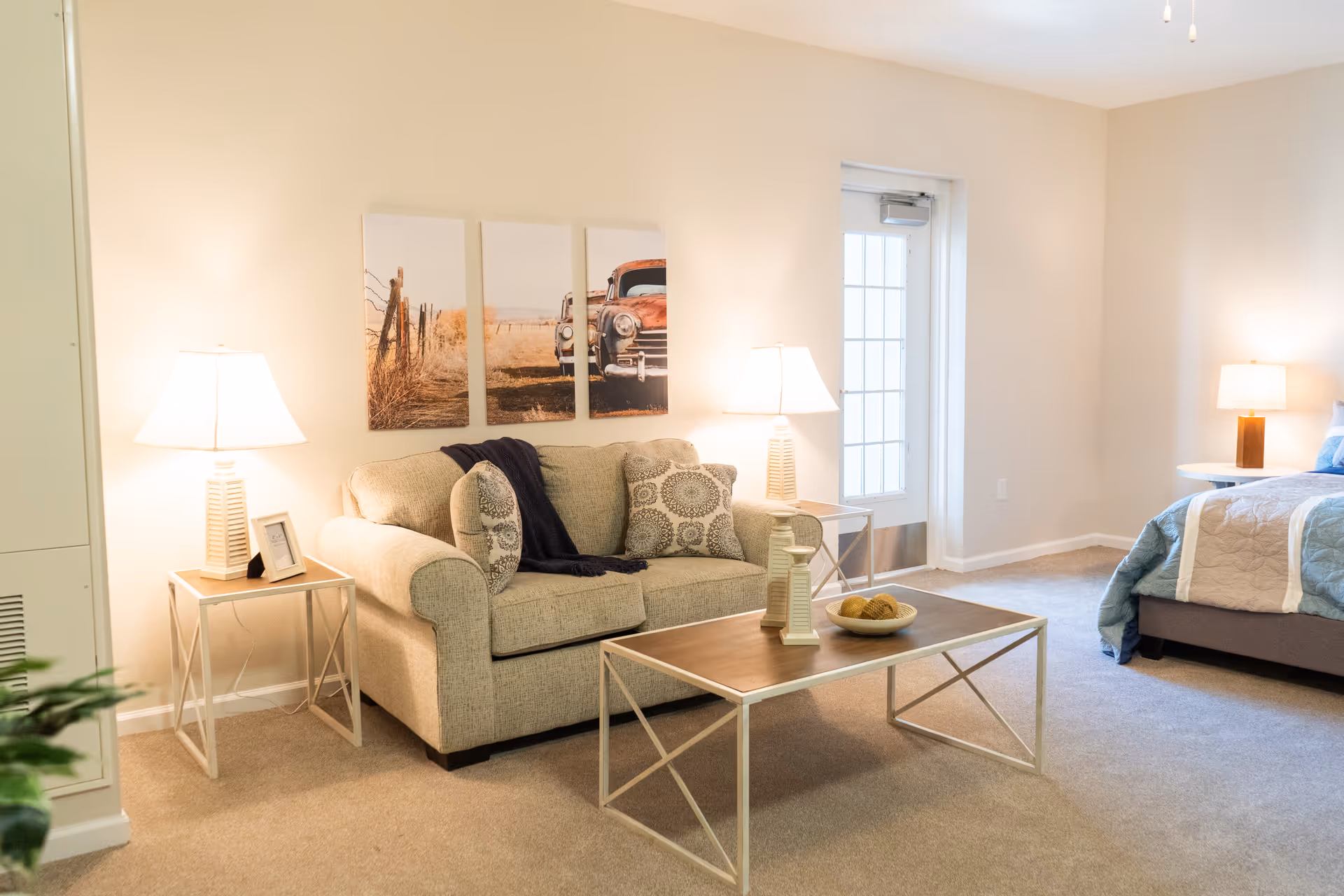 Bright studio living area with a beige sofa, coffee table, side lamps and wall art, with a bed visible in the background.