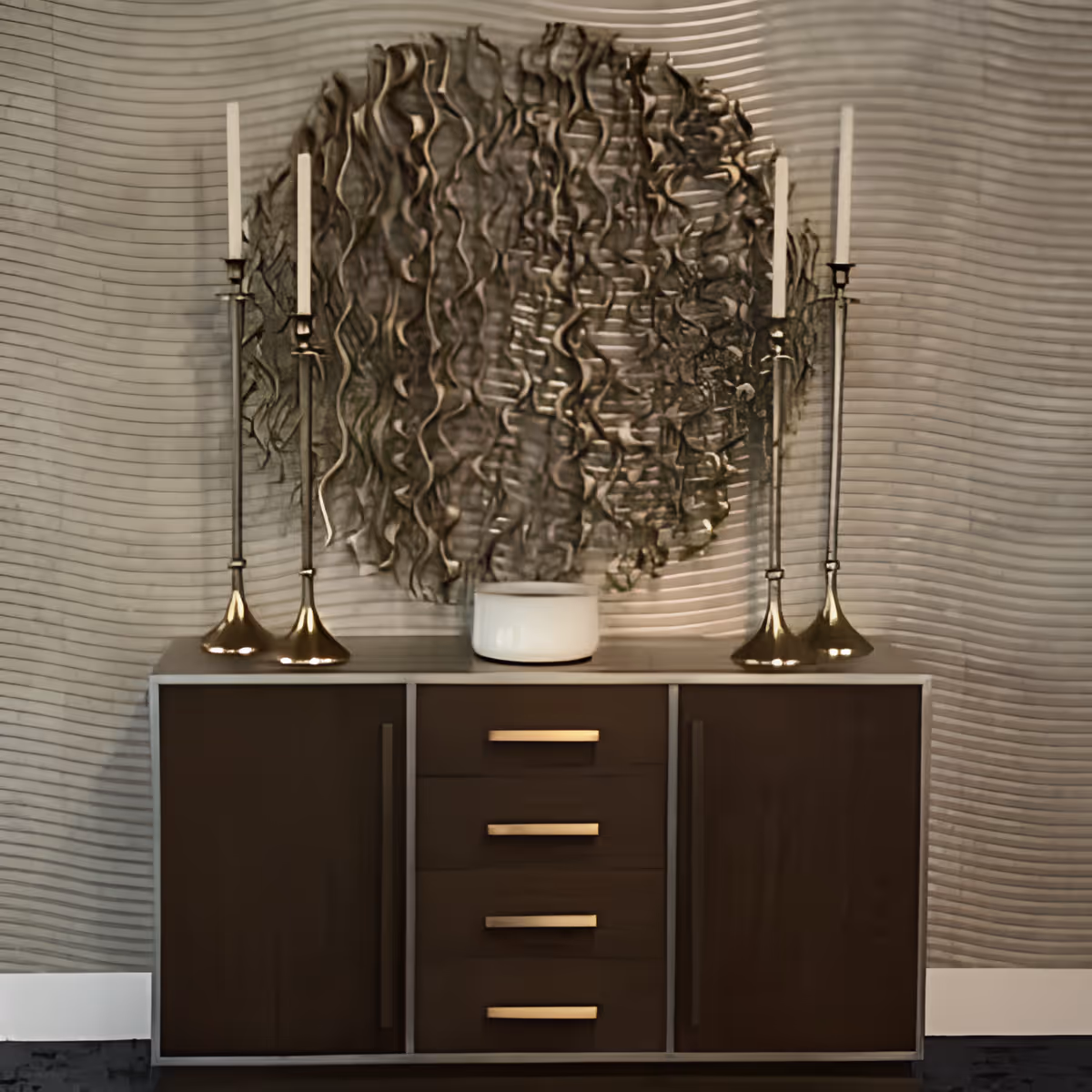 A dark wood console cabinet topped with tall candlesticks and a round abstract metal wall sculpture mounted on textured wallpaper.