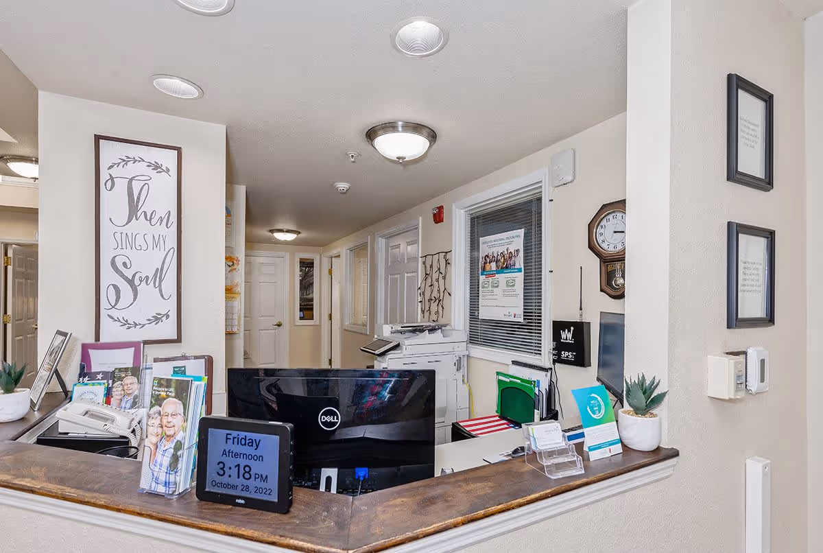 Reception desk area inside Cedar Creek of Seymour facility with a computer monitor, digital clock displaying Friday afternoon 3:18 PM October 28, 2022, framed inspirational wall art saying 'Then sings my soul', office supplies, and a hallway with closed doors in the background.