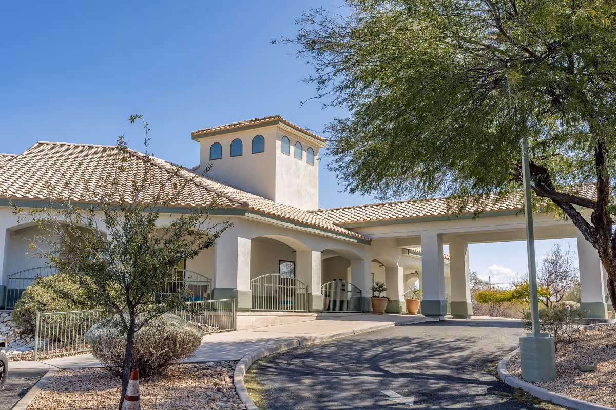 Exterior view of a single-story building with a tiled roof and arched windows, surrounded by desert landscaping including trees and shrubs under a clear blue sky.
