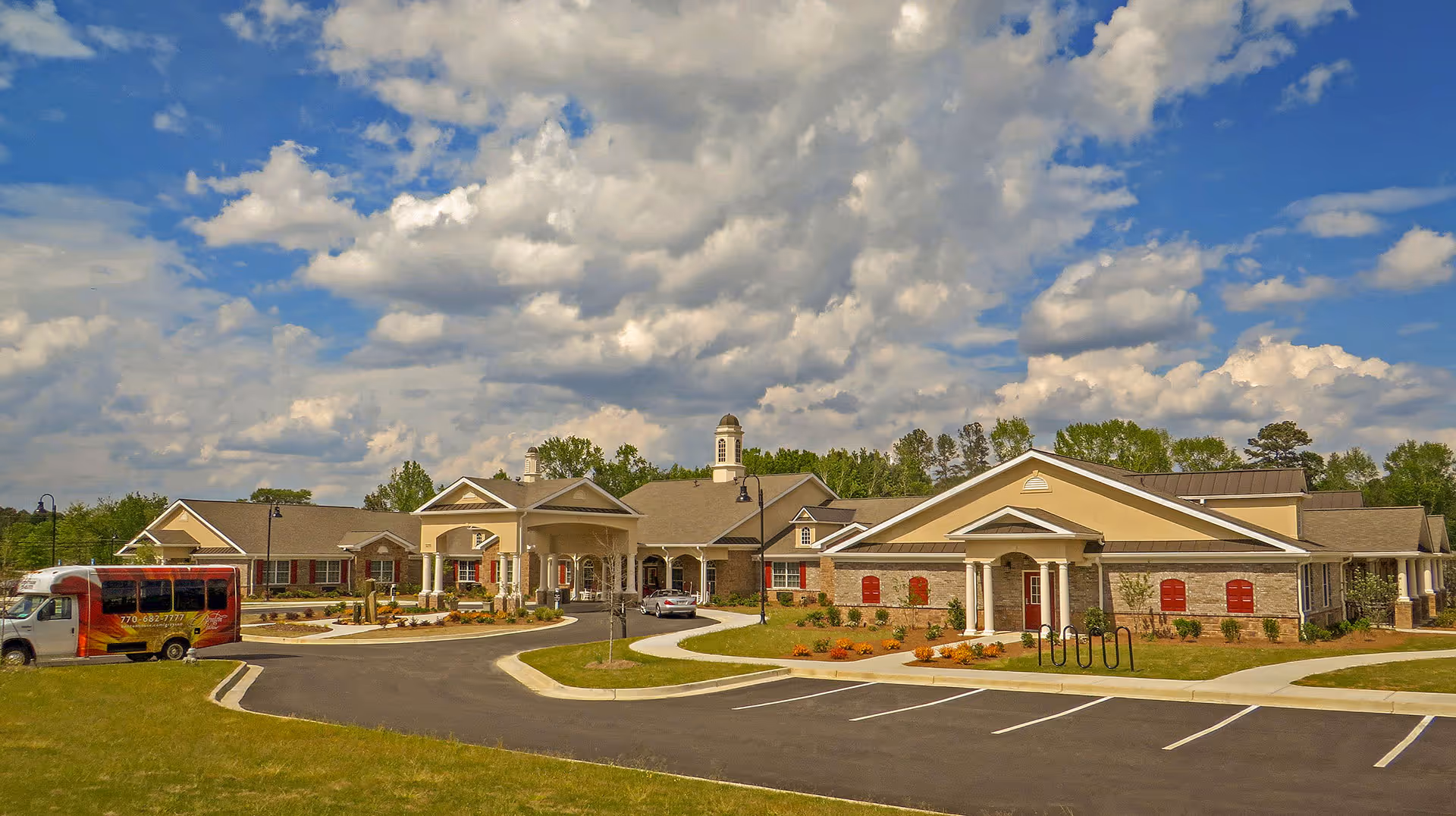 Front exterior view of a single-story senior living facility with a circular driveway, landscaping, and a shuttle bus parked to the left under a partly cloudy sky.