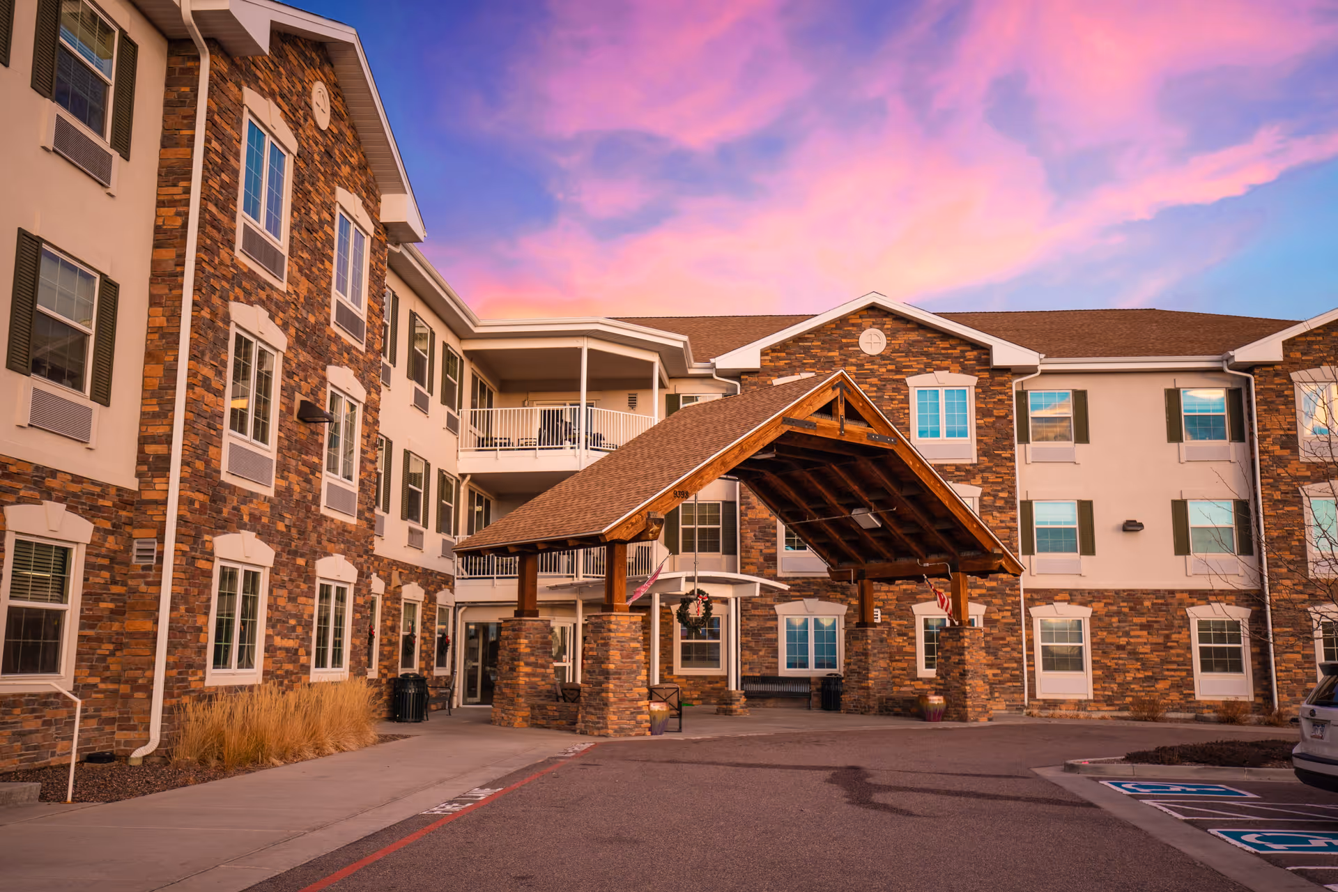 Exterior view of The Suites Parker senior living facility at sunset, showing a multi-story building with stone and beige siding, multiple windows, a covered entrance with wooden beams, and a parking area with handicap spaces.