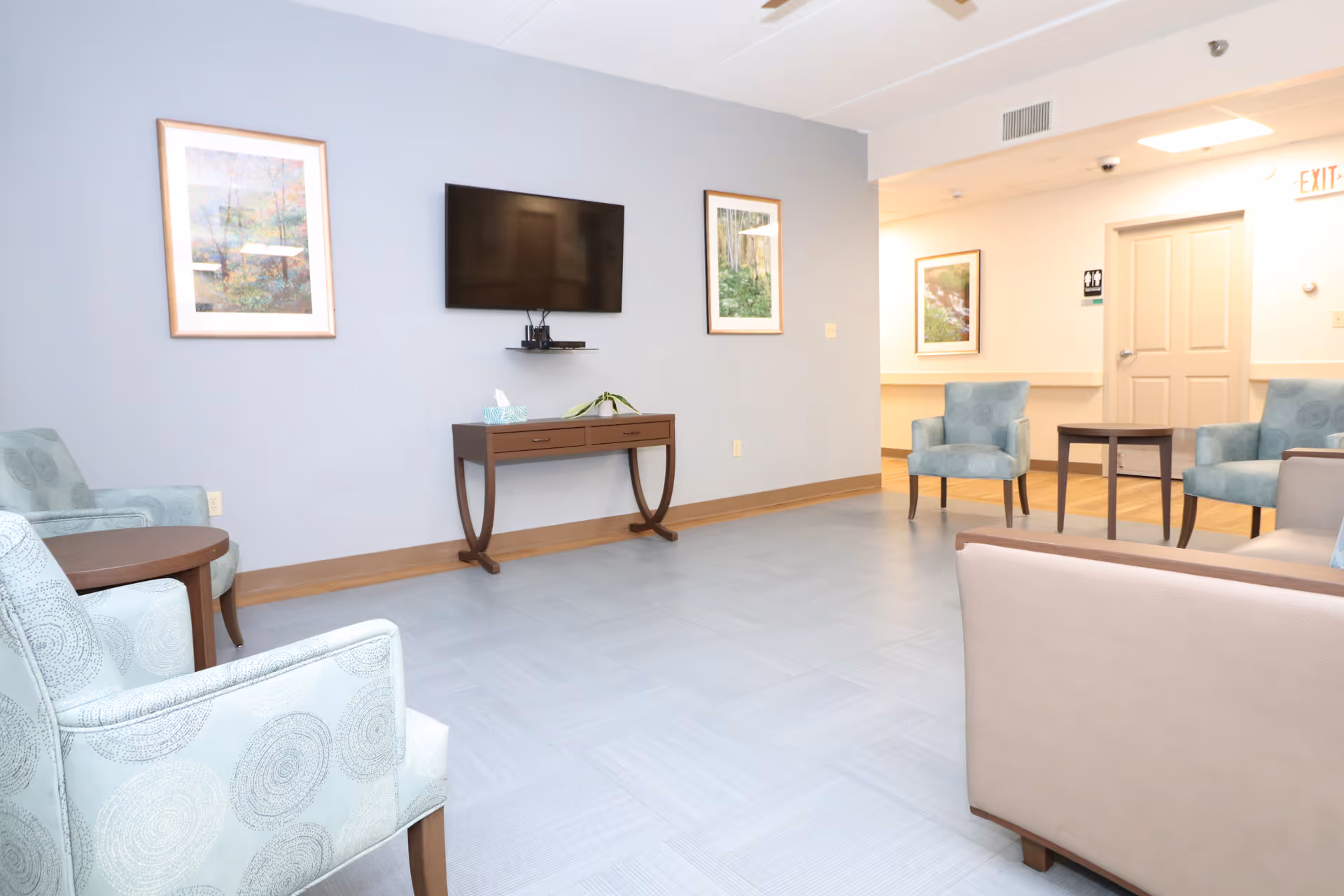 A clean and bright common area with light blue patterned armchairs and a beige sofa arranged around small wooden tables. A flat-screen TV is mounted on a light blue wall above a wooden console table with a tissue box and a small plant. The room has framed nature-themed artwork on the walls and a door with an exit sign in the background.