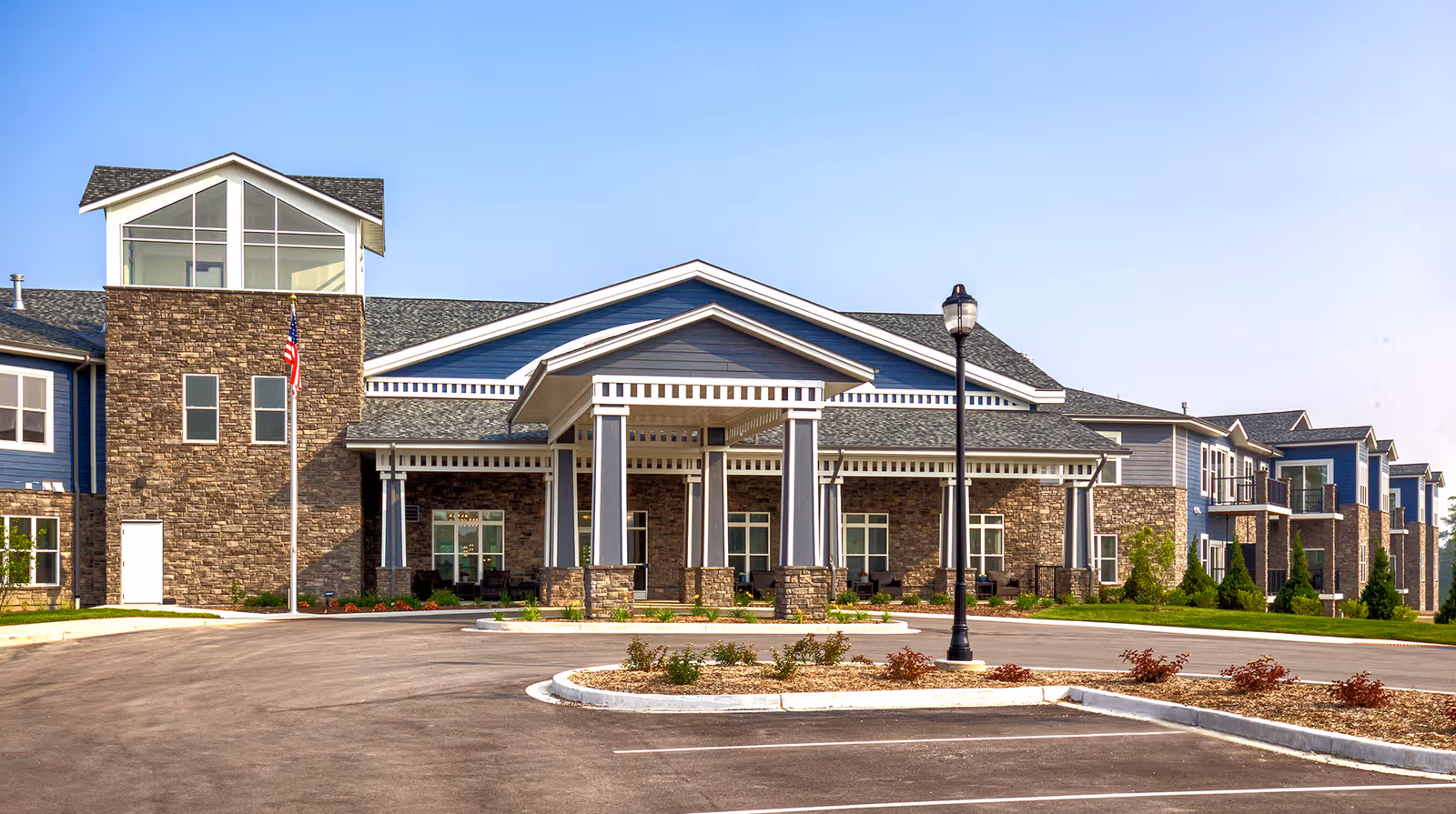Exterior view of a large senior living facility named Stair Crest, featuring a stone and blue siding facade, a covered entrance with columns, an American flag on a flagpole, and a parking area in front under a clear blue sky.