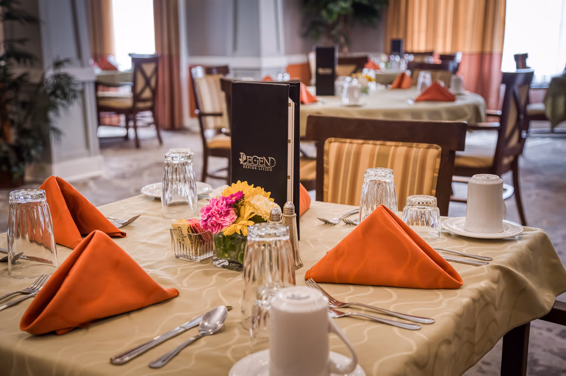 Dining room table set with folded orange napkins, glassware, silverware and a small flower centerpiece in a senior living dining area.