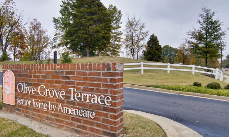 Brick sign for Olive Grove Terrace senior living by Americare at the entrance of the facility, with a curved driveway, grassy area, white fence, and trees in the background under a cloudy sky.