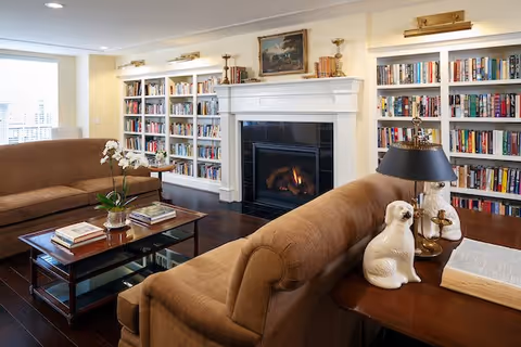 A cozy living room with two brown sofas facing each other, a glass coffee table with books and a plant, a fireplace with a fire burning, and built-in white bookshelves filled with books surrounding the fireplace.