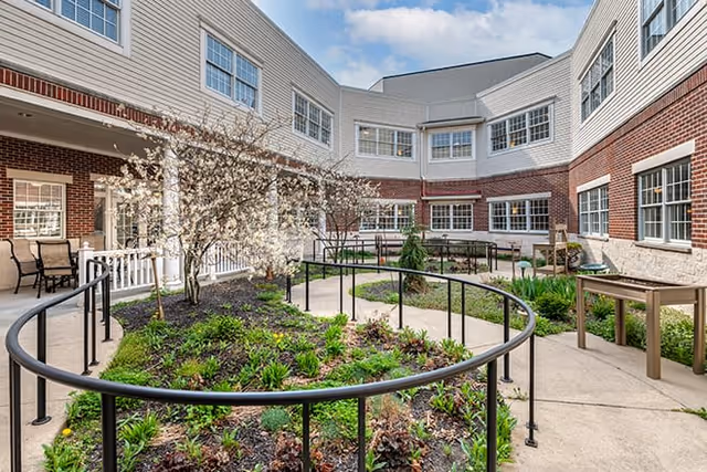 Outdoor courtyard area of a senior living facility with a circular garden bed surrounded by a black metal railing, flowering trees, green plants, and paved walkways. The courtyard is enclosed by a two-story brick building with multiple windows and a covered porch area with chairs.