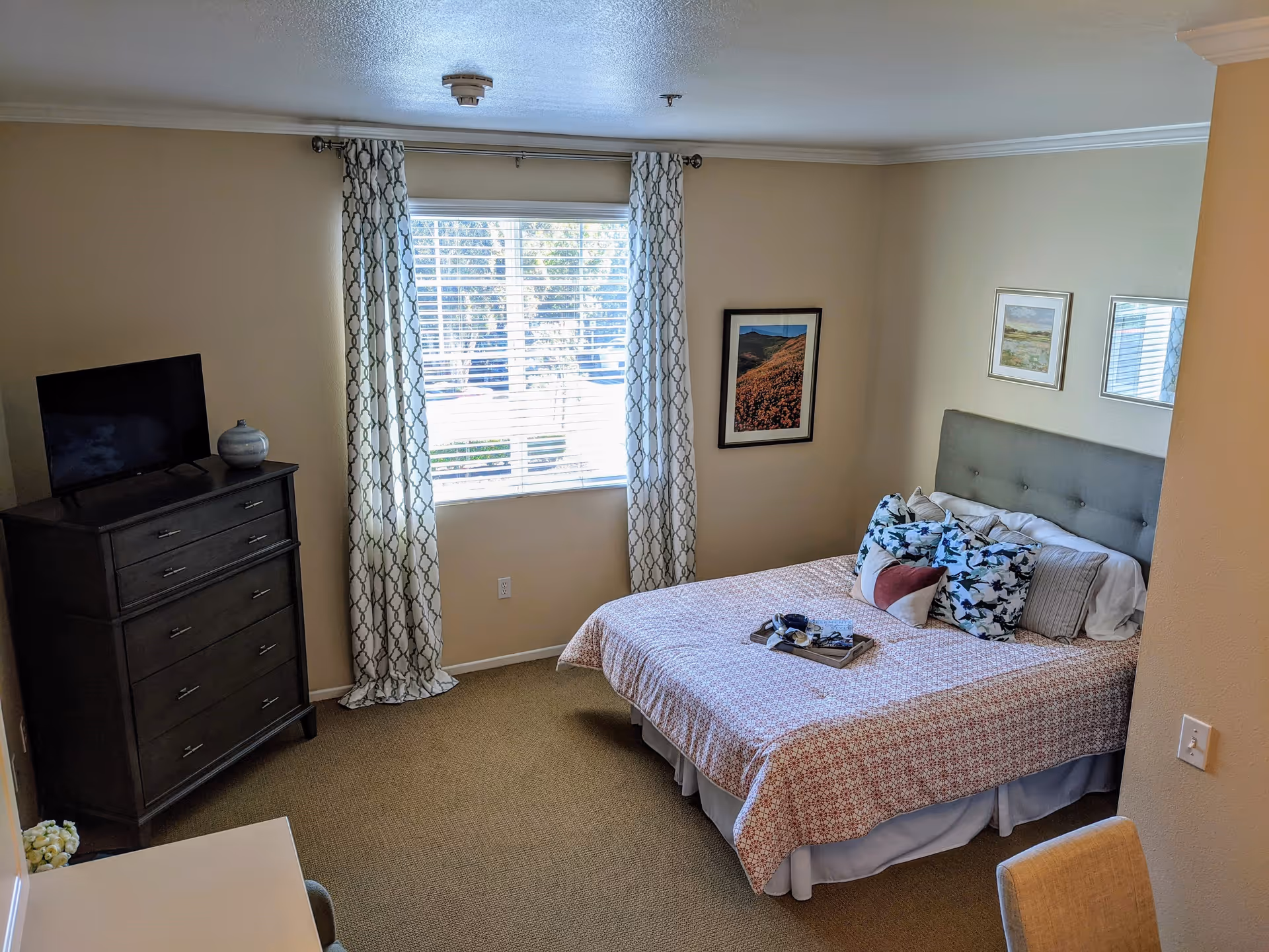 A bedroom with a bed covered in a patterned bedspread and multiple pillows, a window with patterned curtains, a dark wooden dresser with a TV on top, and framed pictures on the walls.