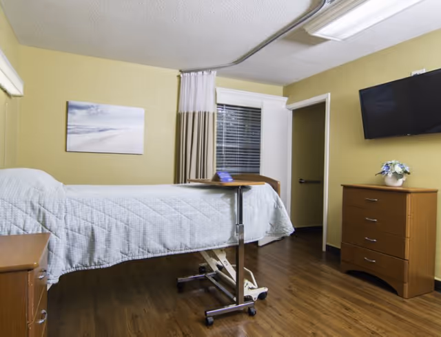 A simple senior care bedroom with a single hospital-style bed, overbed table, dresser, wall-mounted TV, and window.