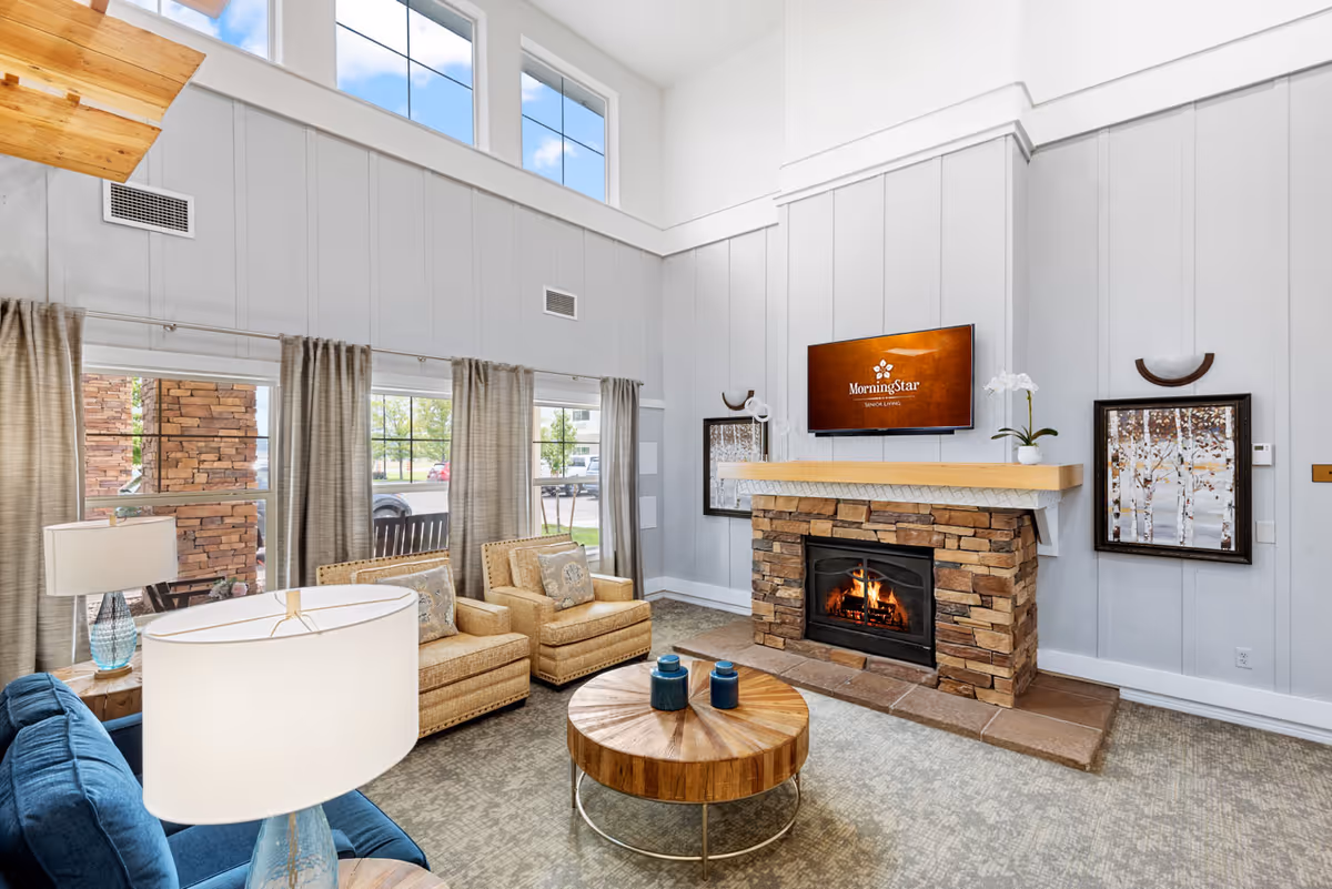 A bright and cozy senior living facility common area with high ceilings and large windows. The room features a stone fireplace with a wooden mantel, above which is a mounted TV displaying the MorningStar Senior Living logo. There are two beige armchairs with patterned cushions, a blue sofa, a round wooden coffee table with decorative items, and a floor lamp with a white shade. Light gray curtains frame the windows, and two framed artworks hang on the walls beside the fireplace.