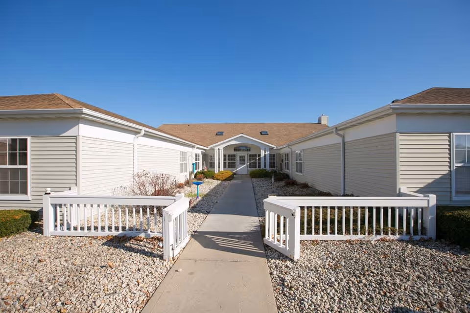 Exterior view of Pleasant Point facility showing a single-story building with beige siding and a brown roof. A concrete walkway leads to the entrance, flanked by white wooden fences and landscaping with rocks and shrubs under a clear blue sky.