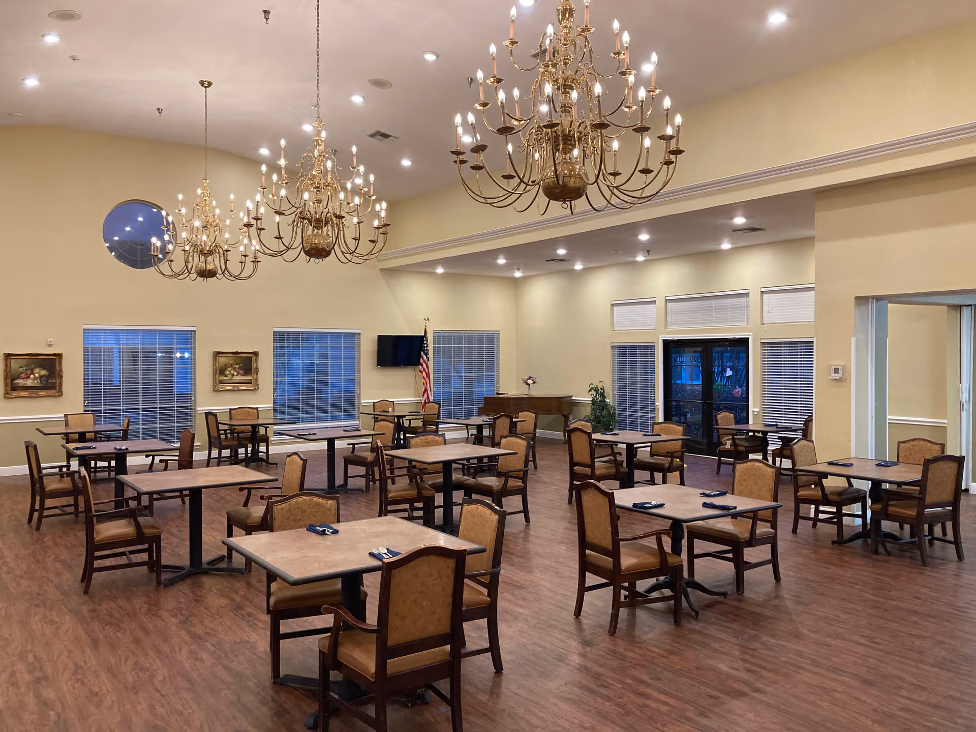 Large dining room with multiple tables and chairs beneath ornate chandeliers and windows.