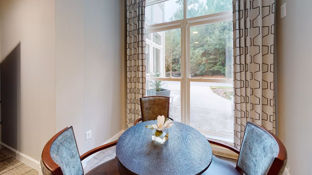 Round dining table with chairs beside a large window with patterned curtains and a small floral centerpiece.