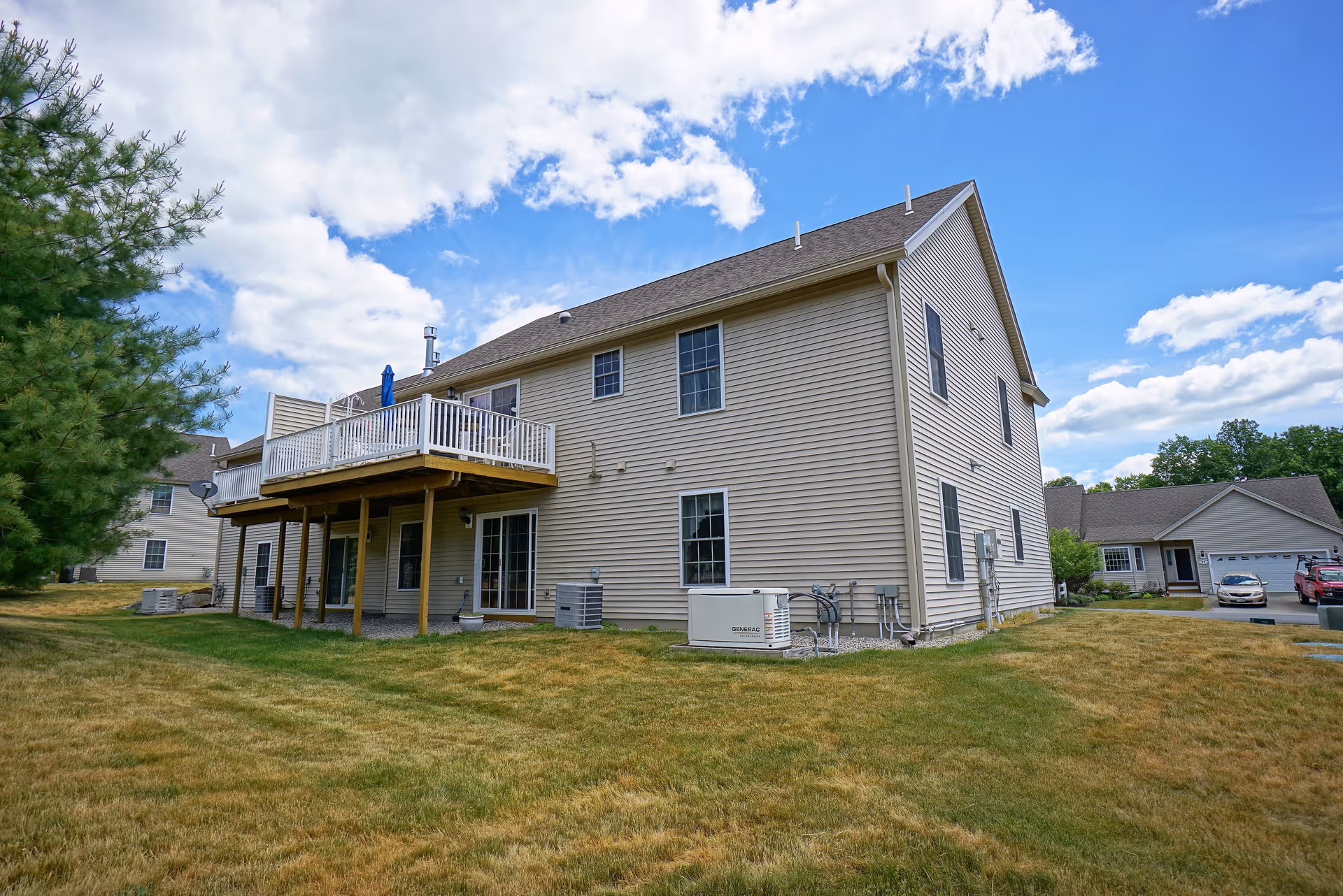 Exterior view of a two-story beige house with a wooden deck on the upper floor, supported by wooden posts. The house has multiple windows and is surrounded by a grassy yard with some trees and neighboring houses visible in the background under a partly cloudy blue sky.