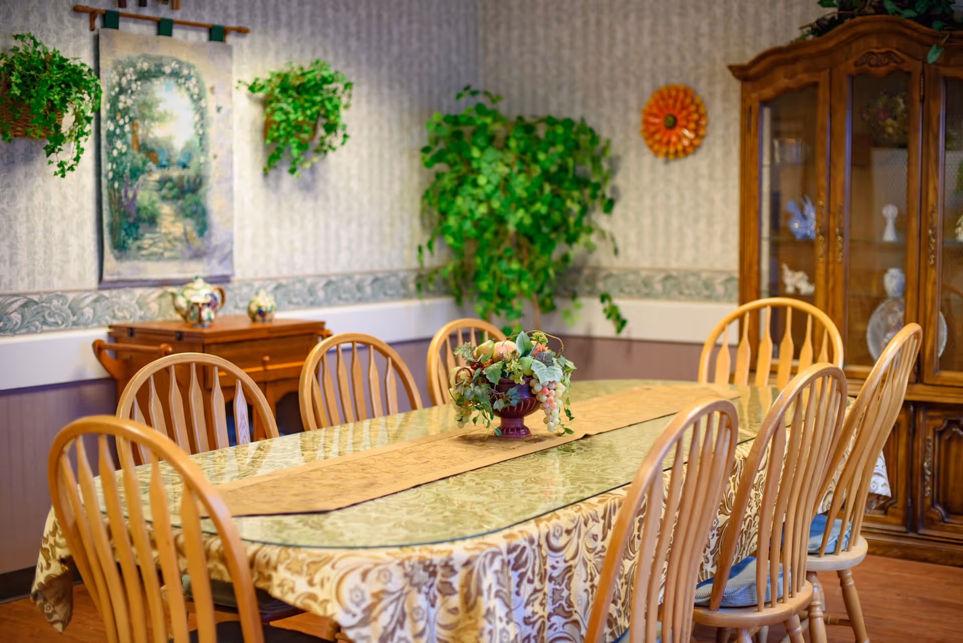 A dining room with a long table covered by a patterned tablecloth and a beige table runner. Eight wooden chairs surround the table. A floral centerpiece is placed in the middle of the table. The room has wallpapered walls with a decorative border, two hanging plants, a framed tapestry, a wooden sideboard, and a glass-fronted wooden cabinet displaying decorative items.
