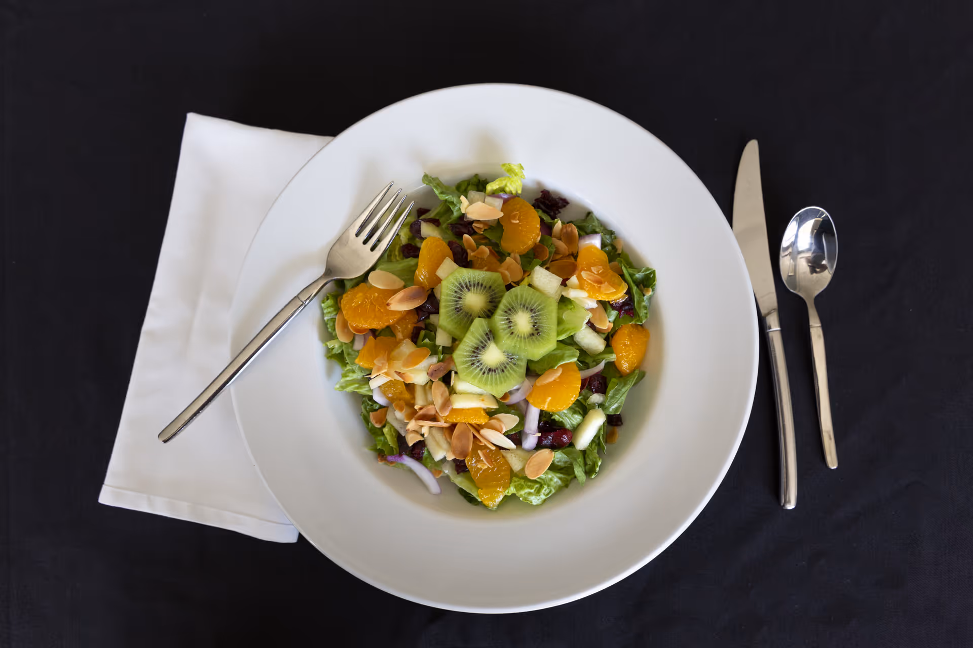 A white plate with a fresh salad containing lettuce, kiwi slices, mandarin orange segments, sliced almonds, red onion, and dried cranberries, placed on a black tablecloth with a white napkin and silver fork, knife, and spoon.