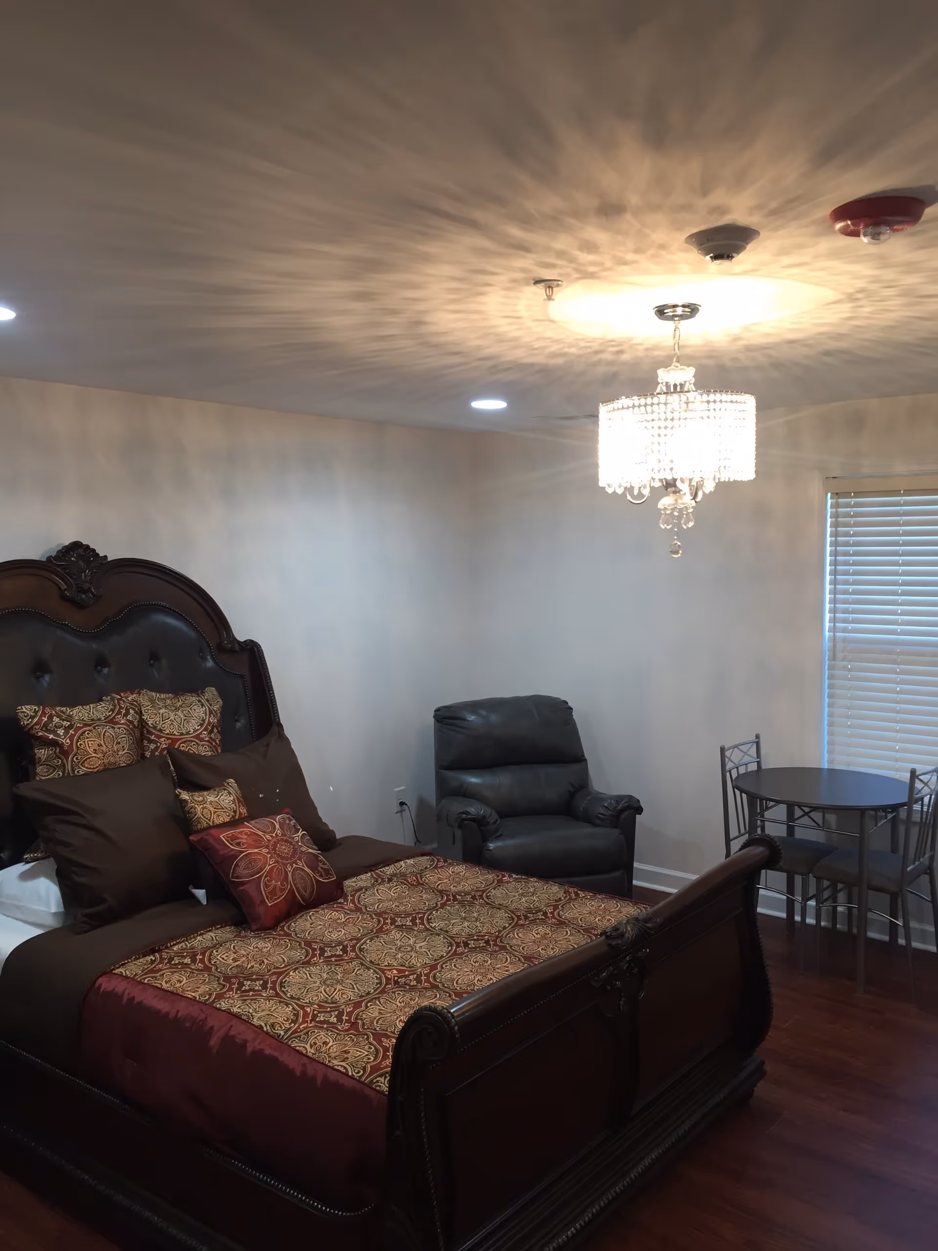 A bedroom with a large ornate wooden bed featuring a patterned bedspread and multiple pillows. There is a black leather recliner chair next to the bed and a small round table with two metal chairs near a window with closed blinds. A crystal chandelier hangs from the ceiling, casting decorative light patterns on the ceiling.