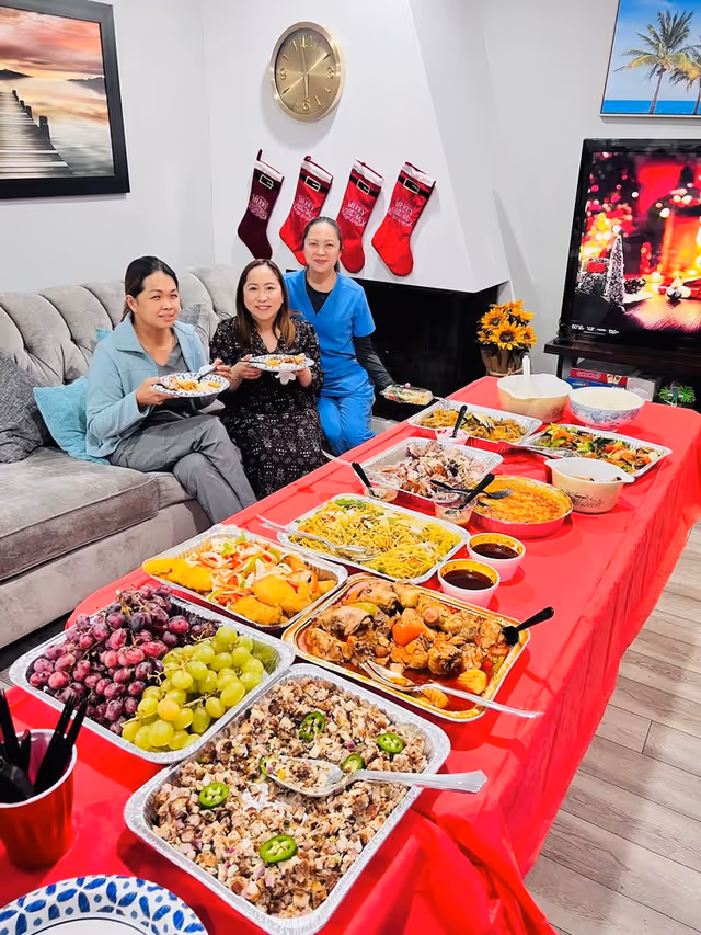 Three women seated on a couch smiling behind a long red-covered buffet table filled with various dishes and fruit in a living room decorated with stockings.
