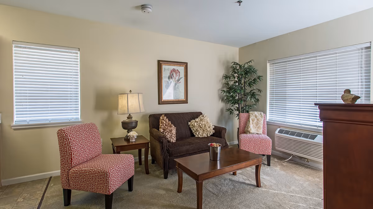 A cozy living room with two patterned red and white chairs, a dark brown loveseat with two decorative pillows, a wooden coffee table with a small bucket of decorative items, a side table with a lamp, a framed floral picture on the wall, a tall green plant in the corner, and two windows with white blinds.