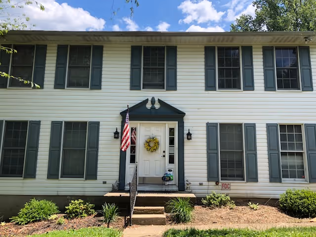Front exterior view of a two-story white building with green shutters and a central entrance. The entrance has a small porch with steps, a white door decorated with a yellow wreath, and an American flag mounted on the left side. There are several windows on both floors and some landscaping with bushes and plants in front of the building under a partly cloudy sky.