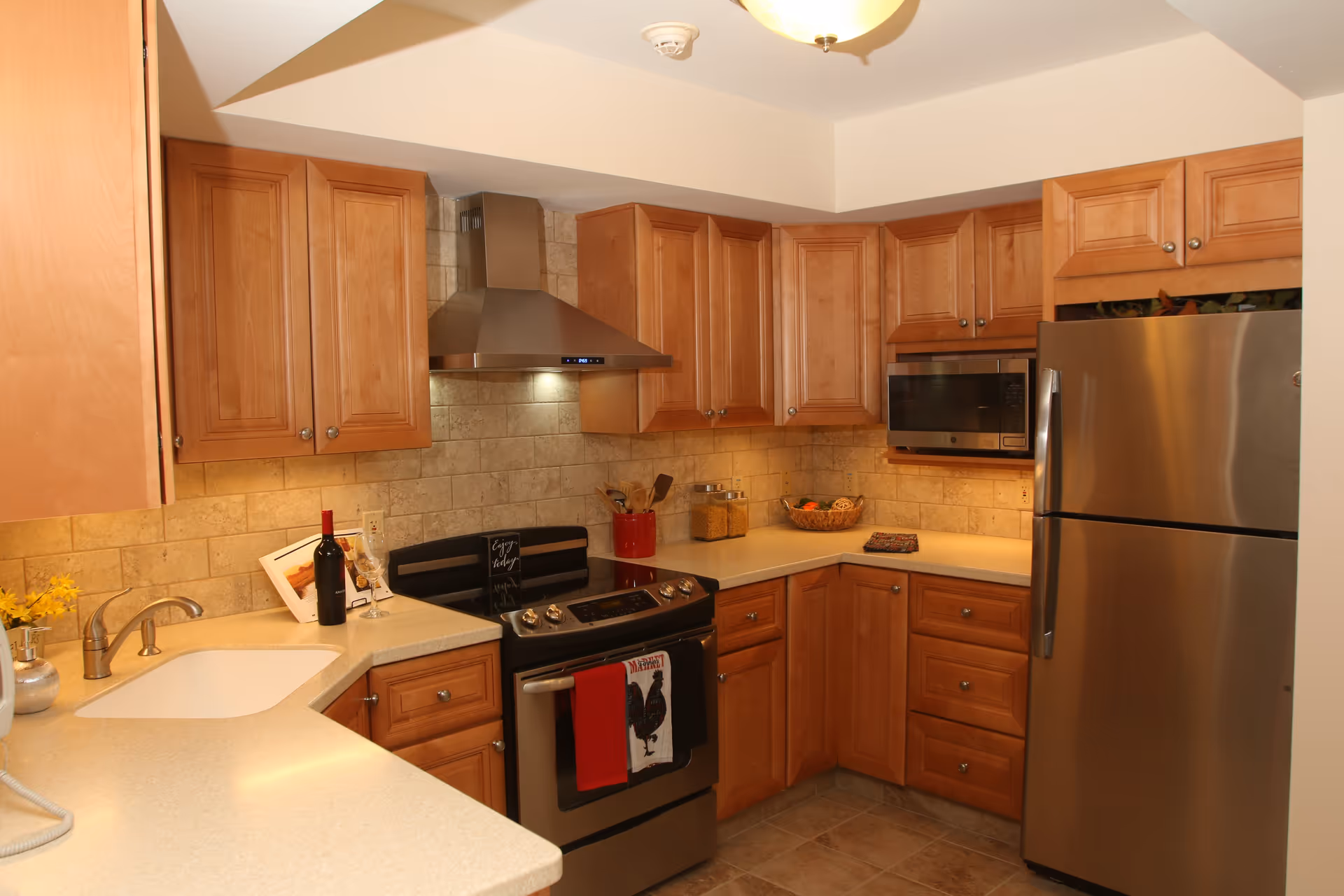 A modern kitchen with wooden cabinets, a stainless steel refrigerator, stove with a range hood, microwave, and beige countertops. There is a sink with a faucet on the left side, a bottle of wine and a wine glass on the counter, and a basket of fruit on the corner counter.
