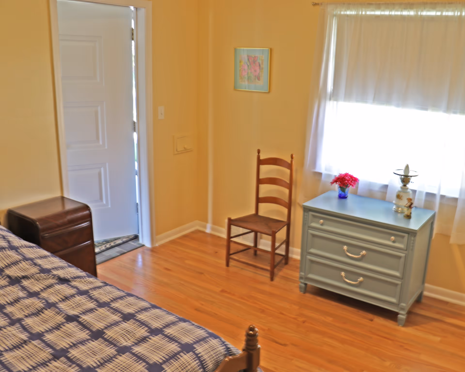 A cozy bedroom with a wooden floor, a bed with a blue and white patterned bedspread, a wooden chair, a light blue dresser with two drawers, a small vase with red flowers, a decorative lamp, a window with sheer white curtains, and a white door slightly open.